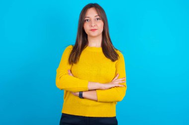 Kendine güveni tam, sakin, sarı örgü süveter giyen, kolları bağlı genç bir Arap kadın. Profesyonel titreşimlerin kendine güvenen pozlarda durduğunu gösterir.