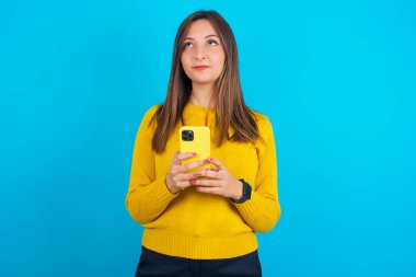 Mavi sırt çantasının üzerine sarı örülü süveter giyen genç Arap kadın cep telefonunu yüksek hızlı internet kullanıyor ve sosyal ağlar internet iletişimine sahip. Modern teknoloji kavramı