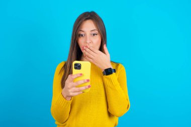 Sarı örgü süveter giyen genç bir Arap kadın mavi arka planda derinden şaşırıyor, akıllı telefon ekranına bakıyor, web sitesinde şok edici haberler okuyor, Omg, bu korkunç.!