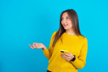 Mutlu, mutlu, sarı örgü süveter giyen genç bir Arap kadın avucunu kaldırıyor ve cep telefonunu cep telefonuyla mesajlaşma ya da video görüşmeleri için kullanıyor.