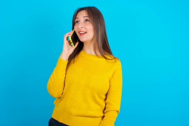 Mutlu görünümlü, sarı örgü süveter giyen genç bir Arap kadın güzel bir telefon konuşması yapıyor ve bir kenara bakıyor, cep telefonuyla konuşurken keyifli ve gülümsüyor.