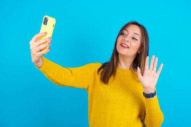Mavi sırt çantasının üzerine sarı örülü süveter giyen genç Arap kadın modern cep telefonunu tutuyor ve merhaba jesti olarak video çağrısı yapıyor. İnsanlar modern teknoloji kavramı