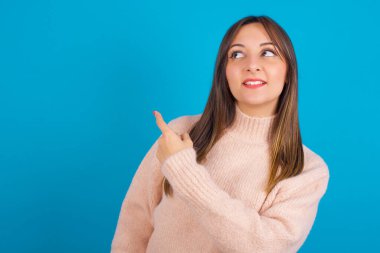 Mavi arka bahçenin üzerine örülmüş süveter giyen genç Arap kadın fotokopi alanını gösteren neşeli, neşeli bir görünüm sergiliyor