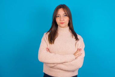 Kendine güveni tam genç bir Arap kadın. Mavi bir kazak giyiyor. Kolları çaprazlanmış kameraya bakıyor.