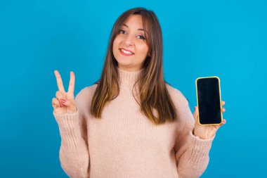 Mavi sırt çantasına örülmüş süveter giyen genç bir Arap kadın. Elinde Ziyaretçi işareti gösteren modern bir cihaz var.