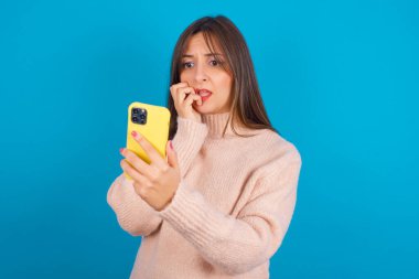 Mavi sırt çantasının üzerine örülmüş süveter giyen, akıllı telefonundan korkunç haberler okuduktan sonra tırnak yiyen genç bir Arap kadının portresi..