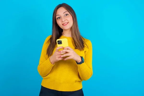 青い背景の上に黄色のニットセーターを着た若いアラブ人女性モックアップコピースペース 携帯電話を使ってSmsメッセージを入力する — ストック写真