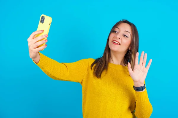 青い背景に黄色のニットセーターを着た若いアラブ人女性は 現代の携帯電話を保持し ハロージェスチャーでビデオ通話波手のひらになります 現代のテクノロジーの概念 — ストック写真