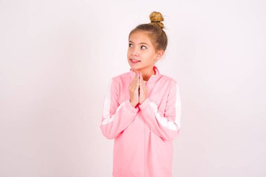 Charming cheerful little caucasian kid girl wearing sport clothing over white background making up plan in mind holding hands together, setting up an idea. Look askance