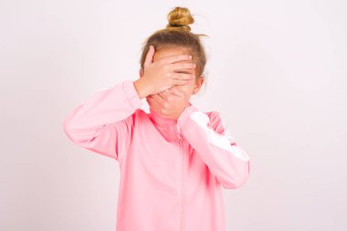 little caucasian kid girl wearing sport clothing over white background Covering eyes and mouth with hands, surprised and shocked. Hiding emotions.