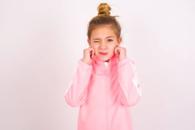 little caucasian kid girl wearing sport clothing over white background covering ears with fingers with annoyed expression for the noise of loud music. Deaf concept.