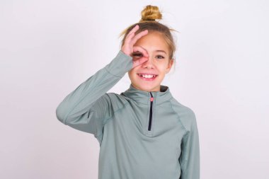 little caucasian kid girl wearing sport clothing over white background doing ok gesture with hand smiling, eye looking through fingers with happy face.