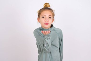 little caucasian kid girl wearing sport clothing over white background looking at the camera blowing a kiss with hand on air being lovely and sexy. Love expression.