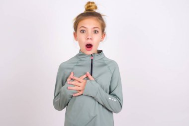 Scared little caucasian kid girl wearing sport clothing over white background looks with frightened expression, keeps hands on chest, being puzzled to notice something strange, People, hush reaction and emotions.