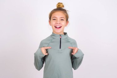 little caucasian kid girl wearing sport clothing over white background points at his body, being in good mood after going shopping and making successful purchases