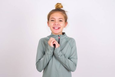 Positive little caucasian kid girl wearing sport clothing over white background  smiles happily, glad to receive pleasant news from interlocutor, keeps hands together. People emotions concept.