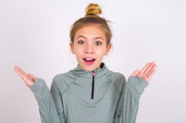 Surprised terrified little caucasian kid girl wearing sport clothing over white background Gestures with uncertainty, stares at camera, puzzled as doesn't know answer on tricky question, People, body language, emotions concept