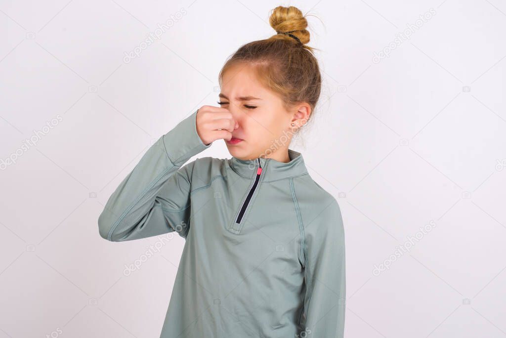 little caucasian kid girl wearing sport clothing over white background smelling something stinky and disgusting, intolerable smell, holding breath with fingers on nose. Bad smell