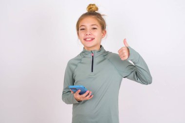 Portrait of little caucasian kid girl wearing sport clothing over white background using and texting with smartphone  happy with big smile doing ok sign, thumb up with fingers, excellent sign