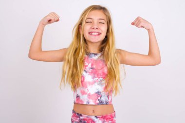 Strong powerful little caucasian kid girl wearing sport clothing over white background toothy smile, raises arms and shows biceps. Look at my muscles!