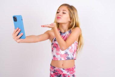 little caucasian kid girl wearing sport clothing over white background blows air kiss at camera of smartphone and takes selfie, sends mwah via online call.