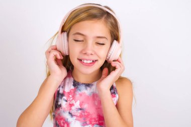 Pleased little caucasian kid girl wearing sport clothing over white background enjoys listening pleasant melody keeps hands on stereo headphones closes eyes. Spending free time with music