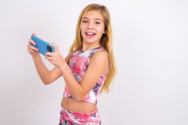 Nice addicted cheerful little caucasian kid girl wearing sport clothing over white background using gadget playing network game