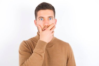 Emotional young caucasian man wearing turtleneck over white background gasps from astonishment, covers opened mouth with palm, looks shocked at camera. Reaction concept