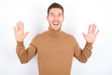 Optimistic young caucasian man wearing turtleneck over white background raises palms from joy, happy to receive awesome present from someone, shouts loudly, Excited model screaming.