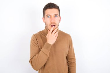 Nervous puzzled young caucasian man wearing turtleneck over white background opens mouth from surprise, reacts on sudden news.