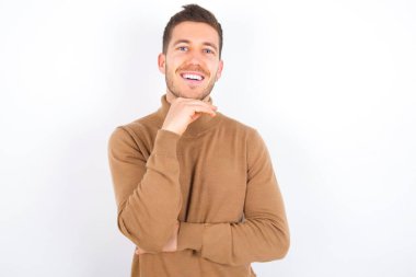 Optimistic young caucasian man wearing turtleneck over white background keeps hands partly crossed and hand under chin, looks at camera with pleasure. Happy emotions concept.