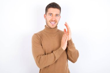 young caucasian man wearing turtleneck over white background clapping and applauding happy and joyful, smiling proud hands together.