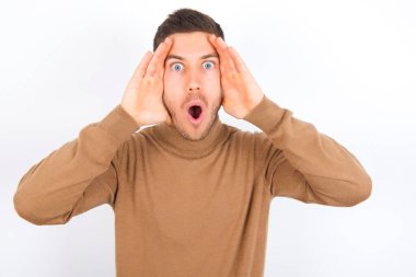 young caucasian man wearing turtleneck over white background with scared expression, keeps hands on head, jaw dropped, has terrific expression. Omg concept