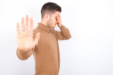 young caucasian man wearing turtleneck over white background covers eyes with palm and doing stop gesture, tries to hide from everybody.