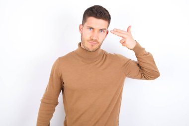 Unhappy young caucasian man wearing turtleneck over white background makes suicide gesture and imitates gun with hand, curves lips, keeps two fingers on temple