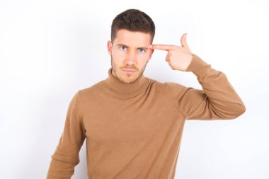 Unhappy young caucasian man wearing turtleneck over white background makes suicide gesture and imitates gun with hand, curves lips keeps two fingers on temple, shoots, being tired of everything,