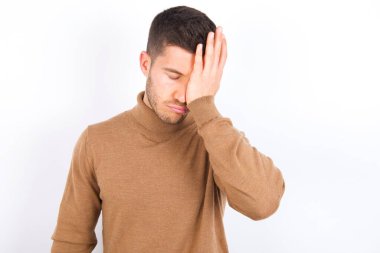 Tired overworked young caucasian man wearing turtleneck over white background has sleepy expression, gloomy look, covers face with hand, has eyes shut, gasps from tiredness, fatigue after party