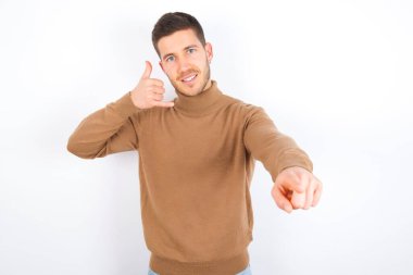young caucasian man wearing turtleneck over white background smiling cheerfully and pointing to camera while making a call you later gesture, talking on phone