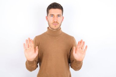 Serious young caucasian man wearing turtleneck over white background pulls palms towards camera, makes stop gesture, asks to control your emotions and not be nervous