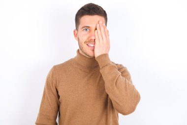 young caucasian man wearing turtleneck over white background covering one eye with her hand, confident smile on face and surprise emotion.