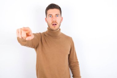 young caucasian man wearing turtleneck over white background Pointing with finger surprised ahead, open mouth amazed expression, something on the front.