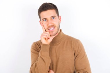 young caucasian man wearing turtleneck over white background looking confident at the camera smiling with crossed arms and hand raised on chin. Thinking positive.