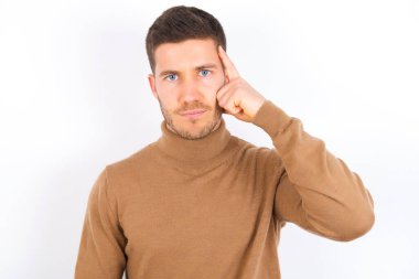young caucasian man wearing turtleneck over white background tries to memorize something, keeps fore finger on temple, reminds information for exam,