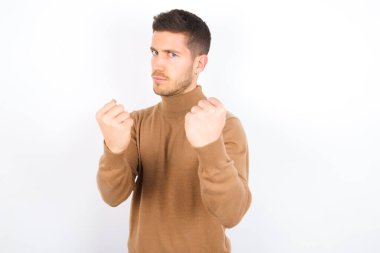 young caucasian man wearing turtleneck over white background Ready to fight with fist defense gesture, angry and upset face, afraid of problem.