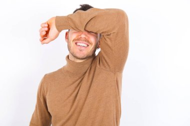young caucasian man wearing turtleneck over white background covering eyes with arm smiling cheerful and funny. Blind concept.