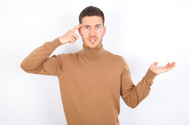 young caucasian man wearing turtleneck over white background confused and annoyed with open palm showing copy space and pointing finger to forehead. Think about it.