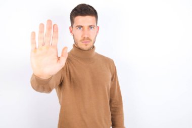 young caucasian man wearing turtleneck over white background doing stop sing with palm of the hand. Warning expression with negative and serious gesture on the face.