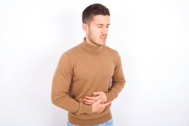 young caucasian man wearing turtleneck over white background with hand on stomach because nausea, painful disease feeling unwell. Ache concept.