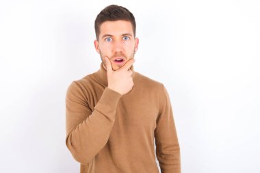 young caucasian man wearing turtleneck over white background Looking fascinated with disbelief, surprise and amazed expression with hands on chin