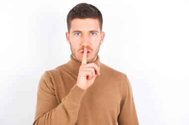 young caucasian man wearing turtleneck over white background makes silence gesture, keeps finger over lips. Silence and secret concept.